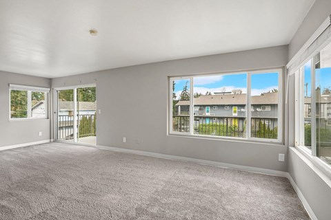 A spacious room with grey carpeting and large windows overlooking a residential area.